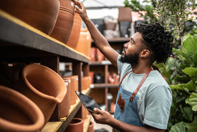 Man assessing different clay pots, dressed in a sage green short sleeved t-shirt and a blue apron with brown leather straps 