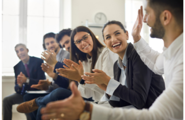 a diverse range of professionals are seated in a row in an office, smiling and clapping in a collaborative manner