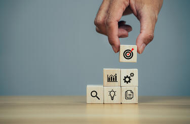 Small building blocks GettyImages-1370817574 Hand using small building blocks each with an image of a lightbulb, magnifying glass, documents, cogs, chart and bullseye