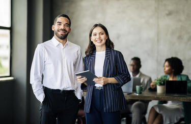 Two colleagues standing together smiling in an office, once is holding a tablet