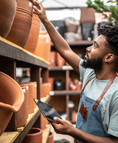 Man assessing different clay pots, dressed in a sage green short sleeved t-shirt and a blue apron with brown leather straps 