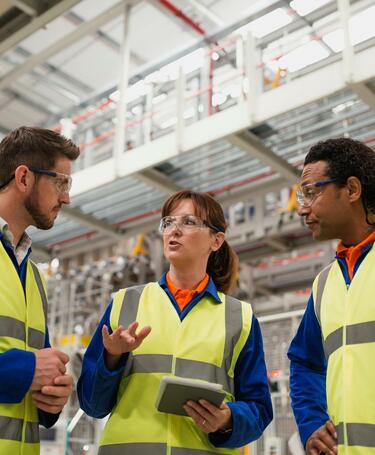A woman and two men in a factory setting, discussing information on an ipad A woman and two men in a factory setting, discussing information on an ipad