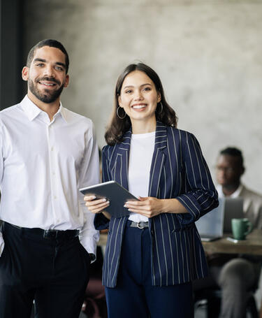 Two colleagues standing together smiling in an office, once is holding a tablet