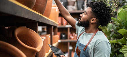 Man assessing different clay pots, dressed in a sage green short sleeved t-shirt and a blue apron with brown leather straps 