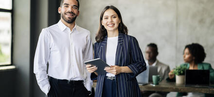 Two colleagues standing together smiling in an office, once is holding a tablet